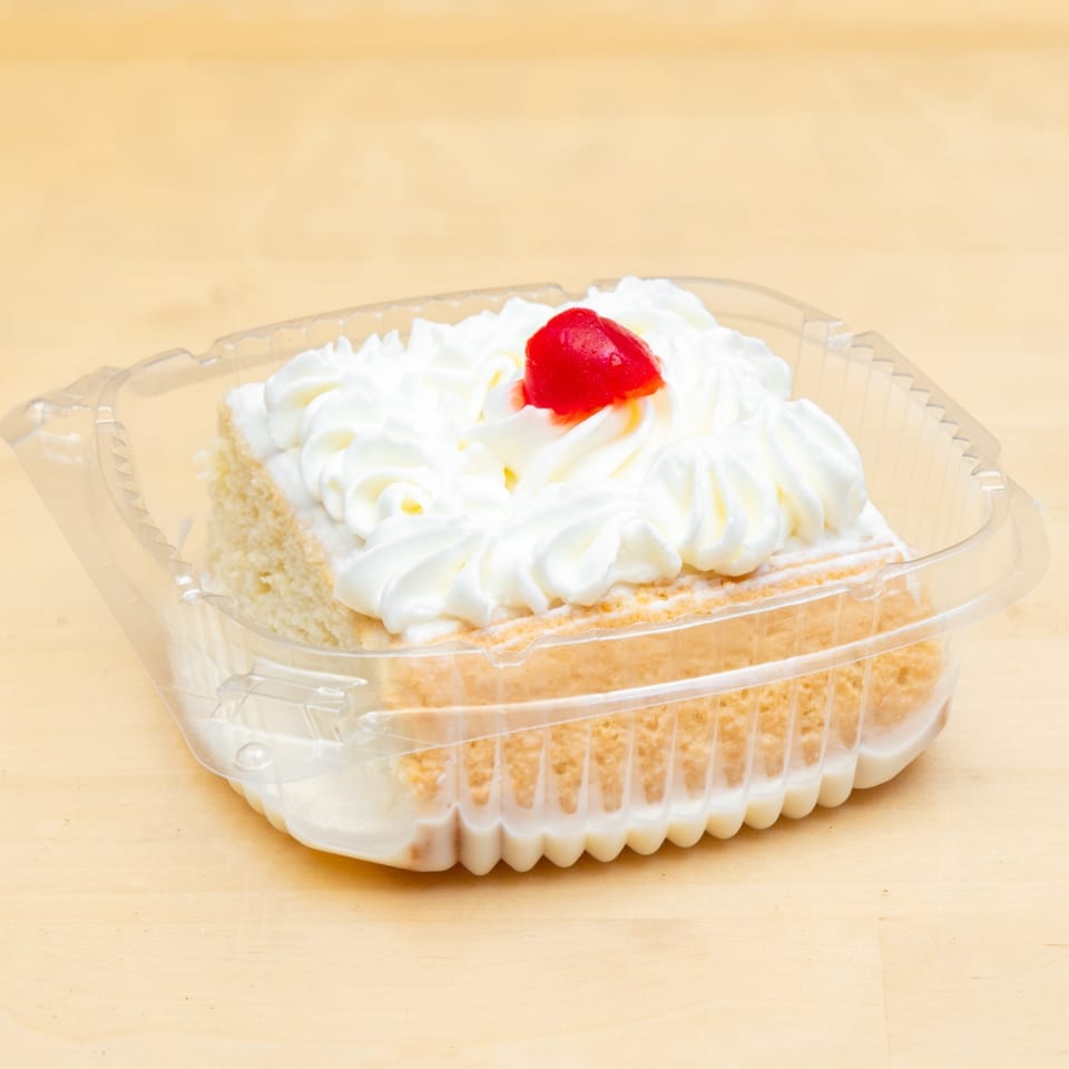 Slice of tres leches cake with whipped cream and a cherry in a clear plastic container.