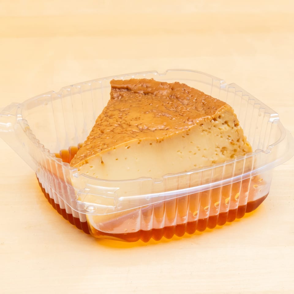 Slice of flan with caramel sauce in a clear plastic container on a light wooden surface.