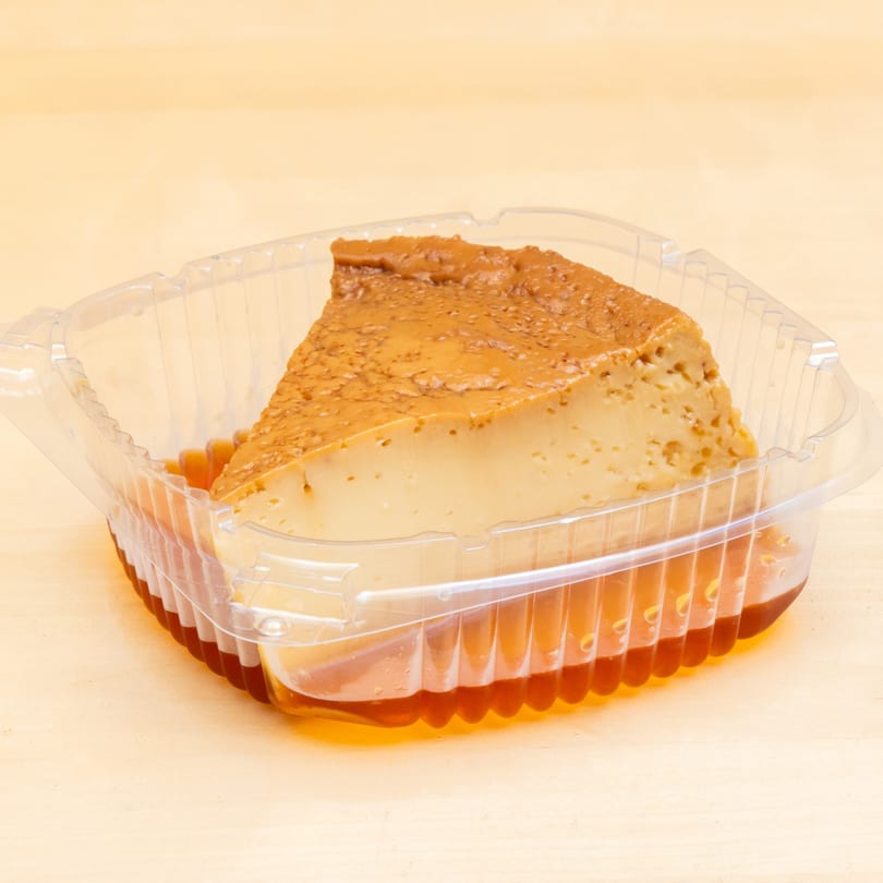 Slice of flan with caramel sauce in a clear plastic container on a light wooden surface.
