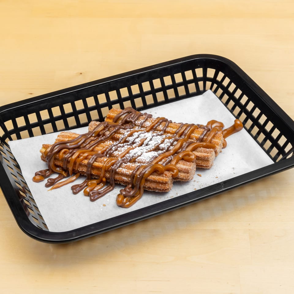 Churros drizzled with chocolate and caramel sauce, dusted with powdered sugar, in a black basket.