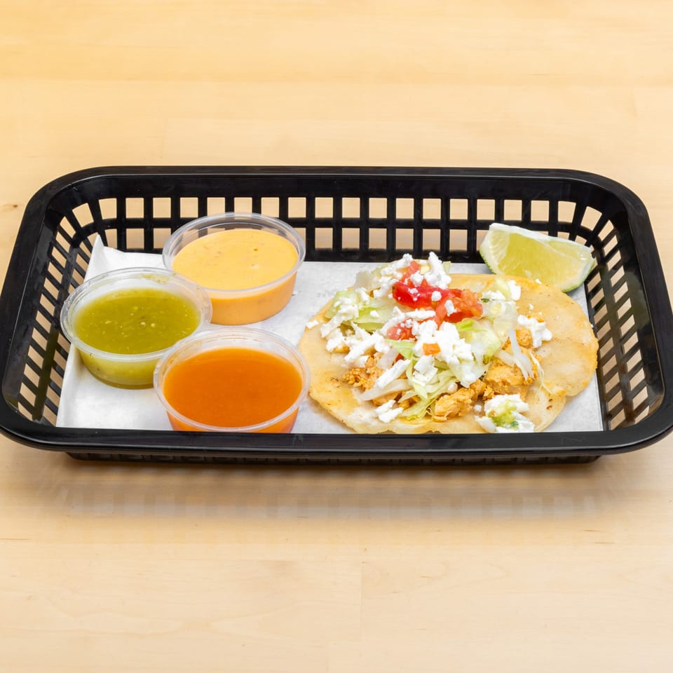 A chicken tostada with lettuce, cheese, and tomato, served with three sauces and a lime wedge.