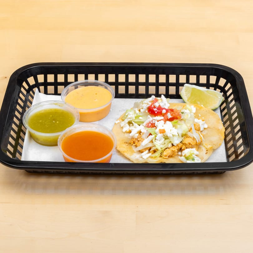 A chicken tostada with lettuce, cheese, and tomato, served with three sauces and a lime wedge.