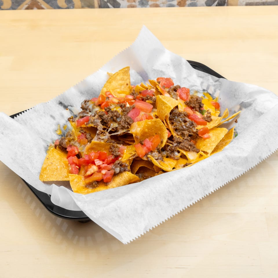 A black basket of nachos with melted cheese, ground beef, and diced tomatoes on a light wooden table.