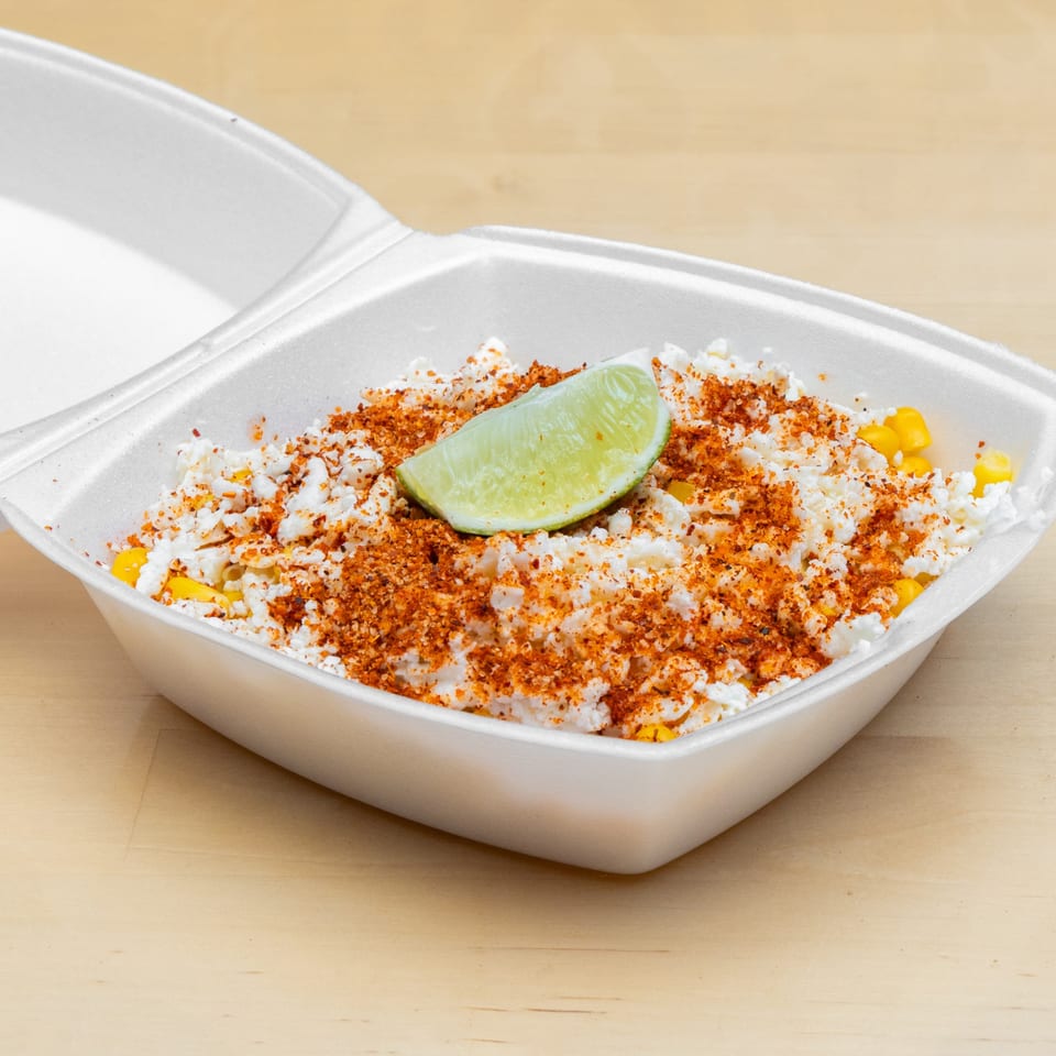 Elote in a white takeout container, topped with cotija cheese, chili powder, and a lime wedge.